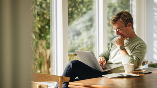  Young man in glasses and a light sweater working happily on a laptop at a wooden table in a bright home office, with large windows overlooking greenery.