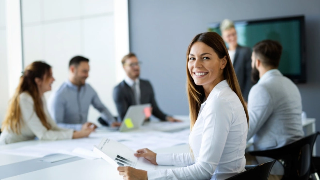 Smiling young woman with brown hair in a white shirt sits at a conference table, holding documents; five blurred colleagues collaborate.